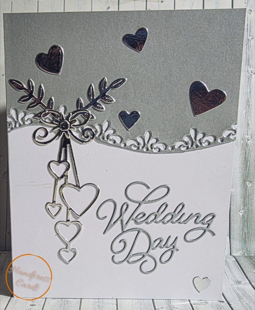 Handmade wedding day card with intricate silver cascading hearts, intertwined rings, botanical sprigs and ornate lace border on a pearl silver and white background.