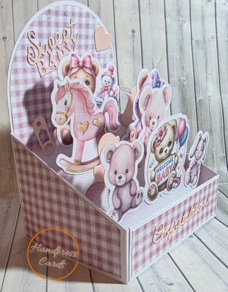 Side view of handmade easel box card showing layered three-dimensional die-cut baby animals and characters in pink and cream tones on a dusky pink gingham background.