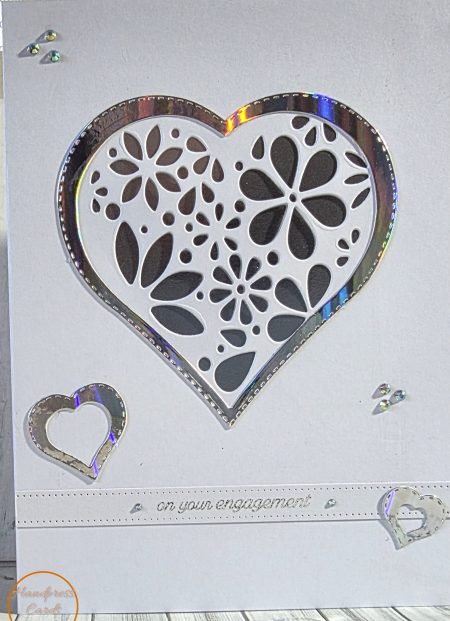 Handmade engagement card featuring a large die-cut floral heart with holographic silver mirror border, small silver heart embellishments, gem details and on your engagement sentiment, on white card stock