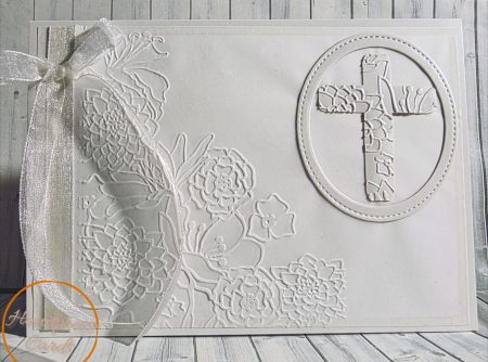 Handmade white christening card featuring deeply embossed floral background with roses and succulents, die-cut floral cross in stitched oval frame and silver organza ribbon bow on white card stock
