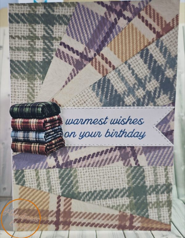 Handmade birthday card with sunburst design made from tartan and houndstooth blanket patterns, featuring a stack of folded plaid blankets and a stitched banner with "warmest wishes on your birthday" in navy script